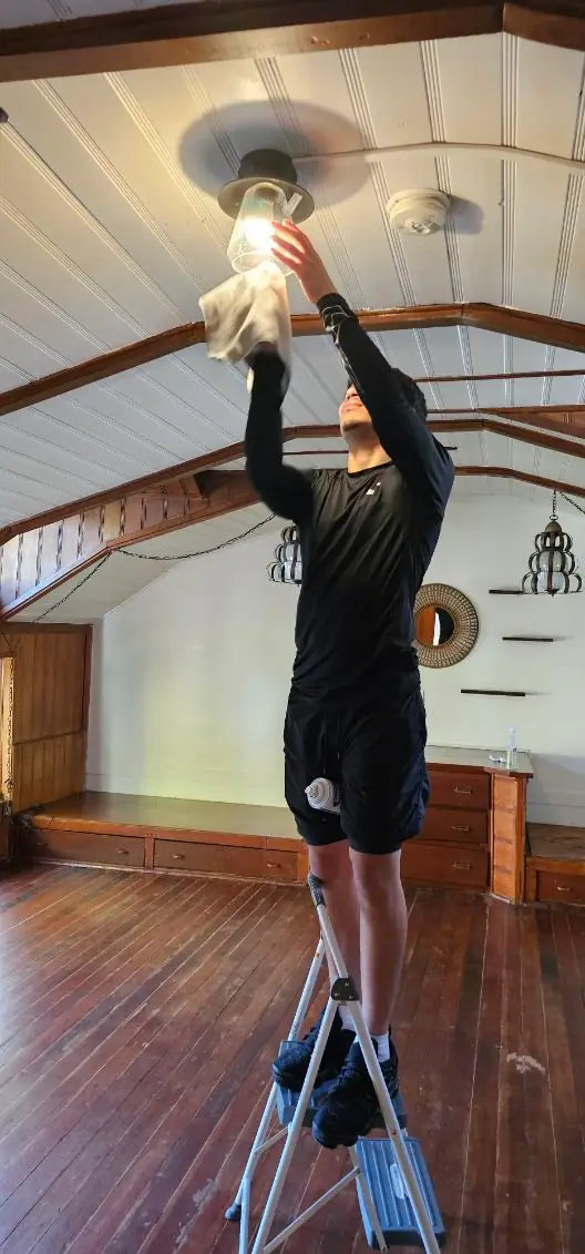 A crew member on a stepladder cleaning a ceiling fan under a vaulted wood ceiling